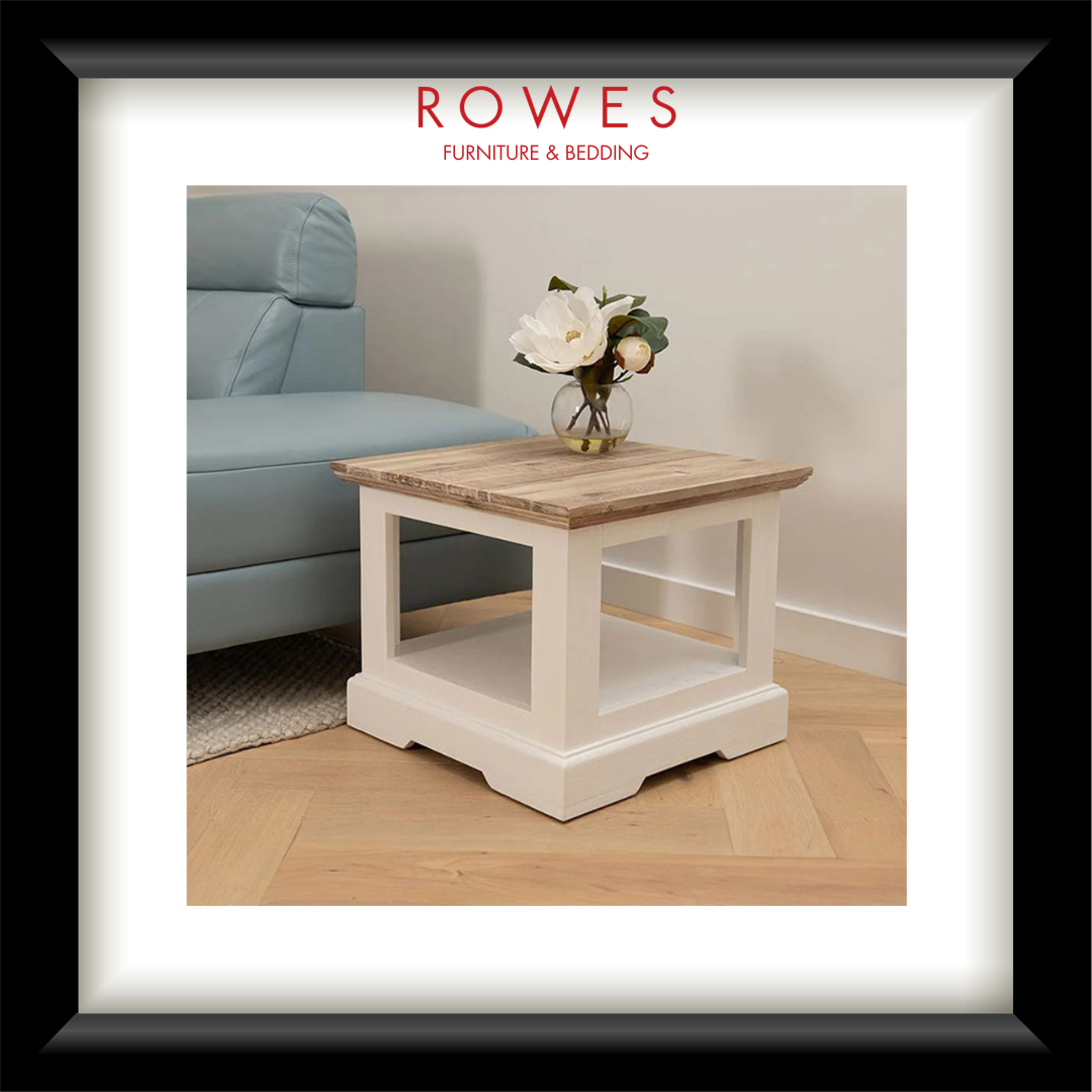 A framed interior photo showing a small square side table with a light natural wood top and white painted frame, placed in a living room beside a pale blue sofa. The frame includes a white mat and a black outer border, with the text 'ROWES FURNITURE & BEDDING' at the top.
