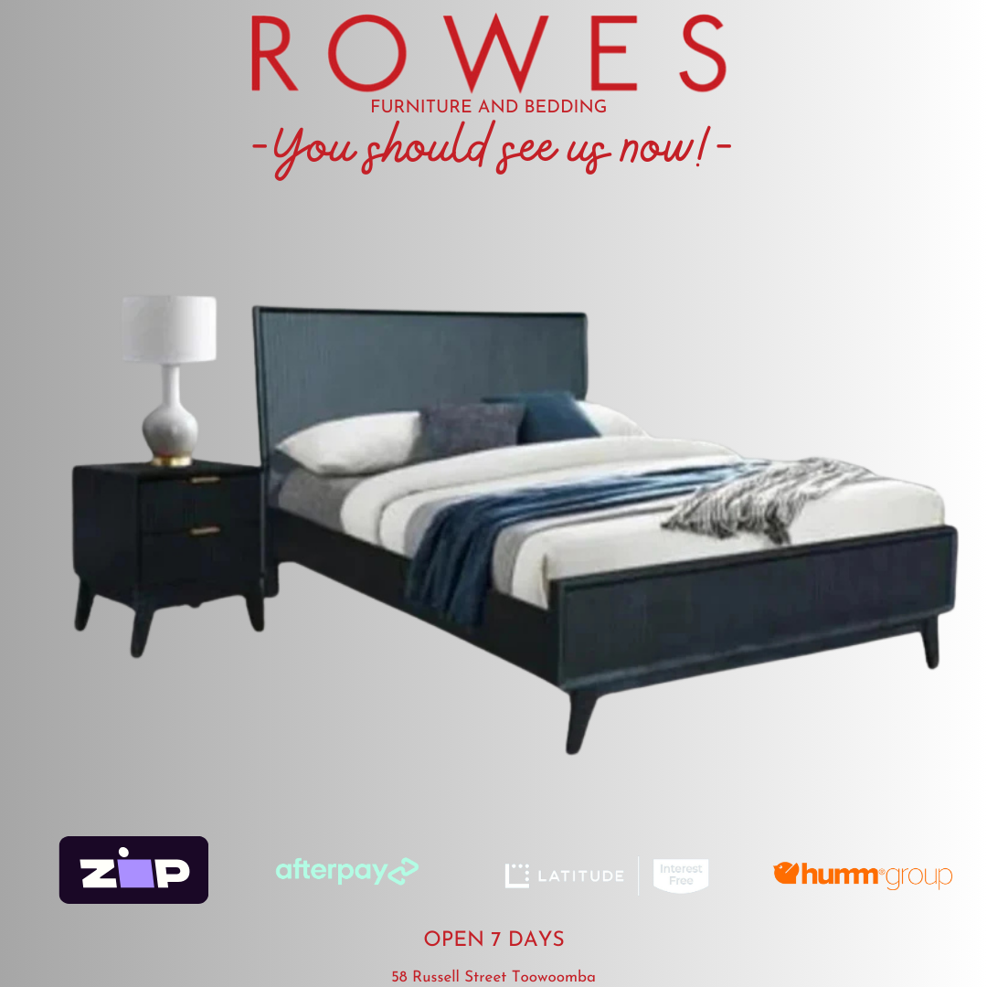 A contemporary bedroom setting featuring a black wooden bed frame with an angled headboard, solid timber drawers, and metal handles, alongside a matching nightstand, dressed with blue bedding. A lamp and decorative items top the nightstand.