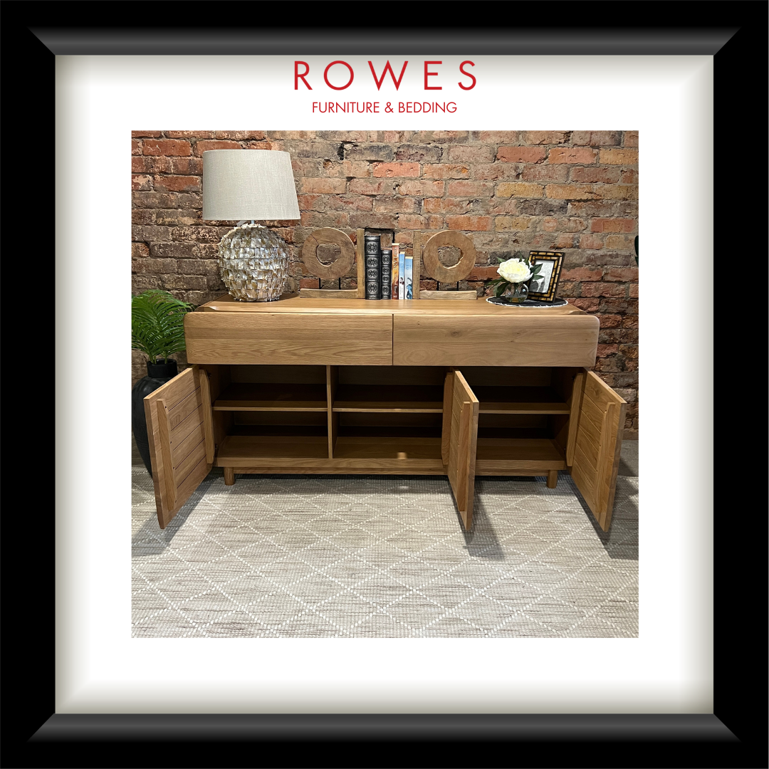 A framed furniture catalog or print featuring a wooden sideboard with open doors, styled with a lamp, books, and decor against a brick wall. The image is matted with a white border and enclosed in a black frame, with the text 'ROWES FURNITURE & BEDDING' at the top.