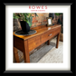 A hardwood hall table on a carpeted floor against a brick wall. On top of the hall table there are some flowers, a photo frame and an ornament. The image features the Rowes branding.