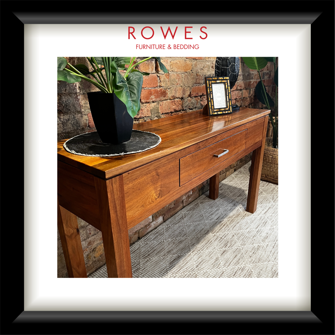 A hardwood hall table on a carpeted floor against a brick wall. On top of the hall table there are some flowers, a photo frame and an ornament. The image features the Rowes branding.