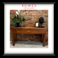 A hardwood hall table on a carpeted floor against a brick wall. On top of the hall table there are some flowers, a photo frame and an ornament. The image features the Rowes branding.