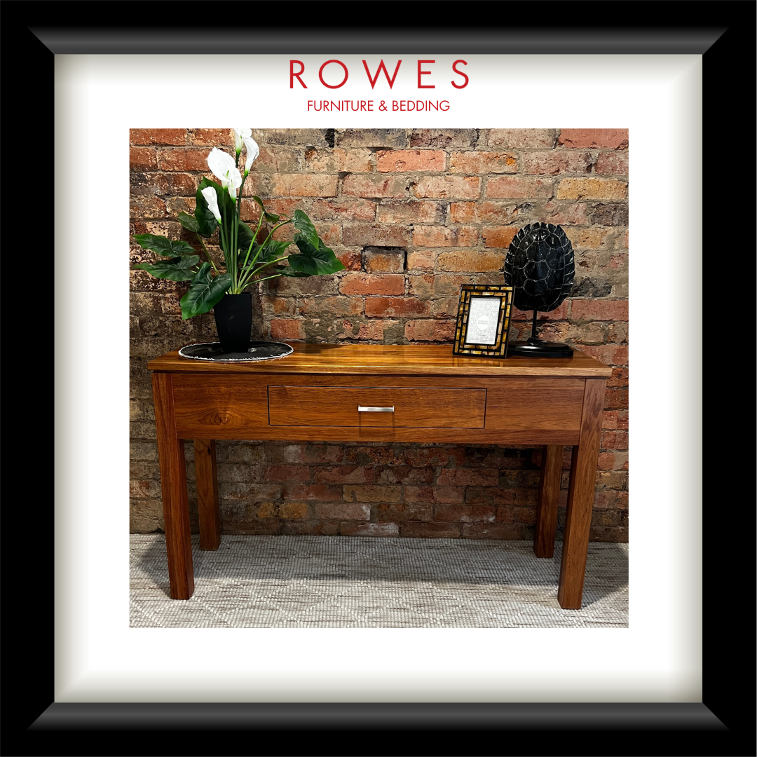 A hardwood hall table on a carpeted floor against a brick wall. On top of the hall table there are some flowers, a photo frame and an ornament. The image features the Rowes branding.
