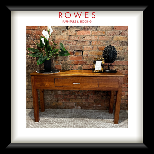A hardwood hall table on a carpeted floor against a brick wall. On top of the hall table there are some flowers, a photo frame and an ornament. The image features the Rowes branding.