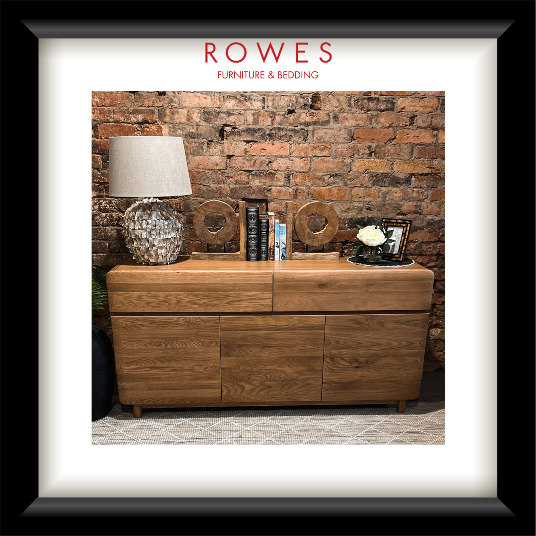 A framed image containing an oak buffet with a lamp and some books on the top surface. The frame has the Rowes branding. 