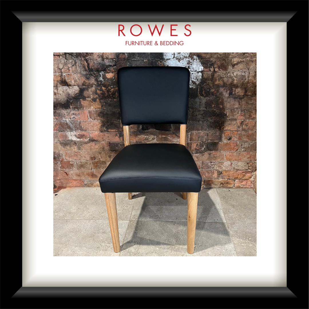 A framed product photograph of a modern dining chair with a black upholstered seat and backrest and light wood legs, set against a brick wall. The photo is matted with a white border and enclosed in a black frame, with the text 'ROWES FURNITURE & BEDDING' printed at the top.