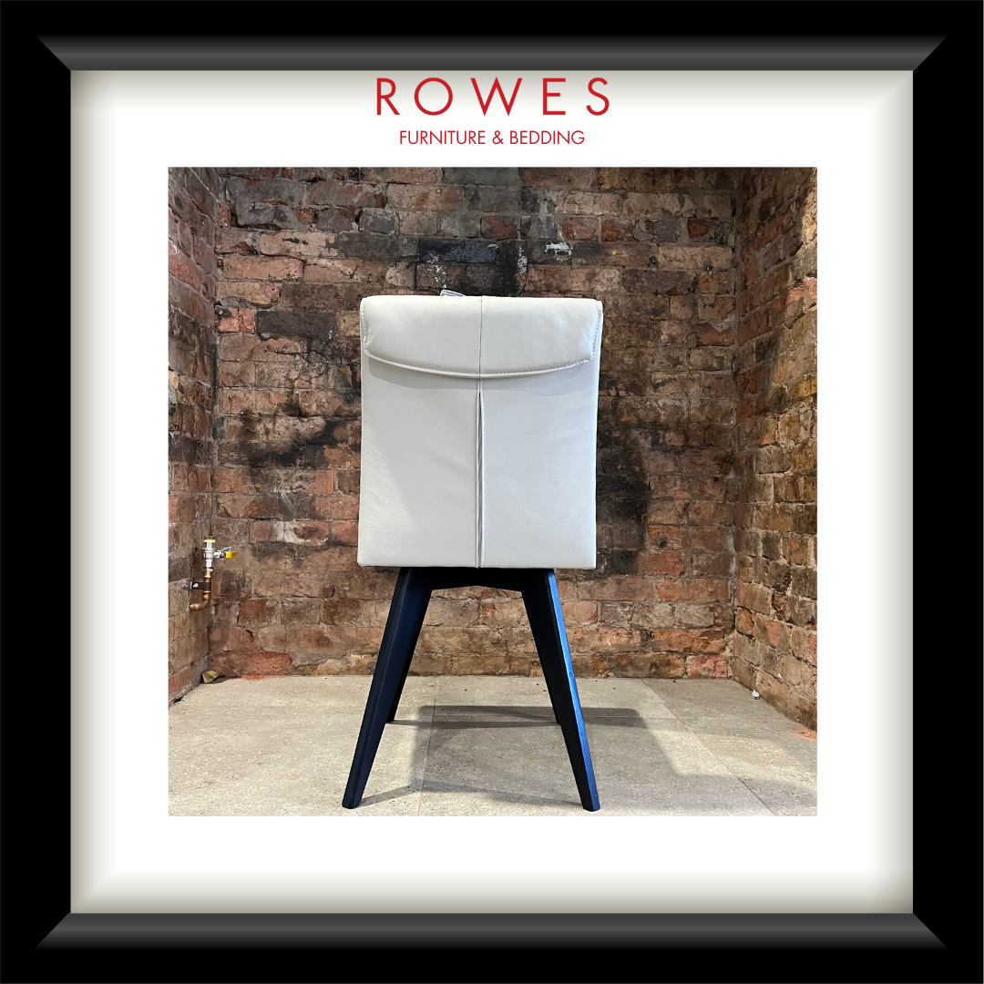 A framed product photograph of a modern dining chair with a white upholstered backrest and dark blue angled legs, set against a rustic brick wall. The image is presented in a white mat and a thick black frame, with the brand name 'ROWES FURNITURE & BEDDING' printed at the top.