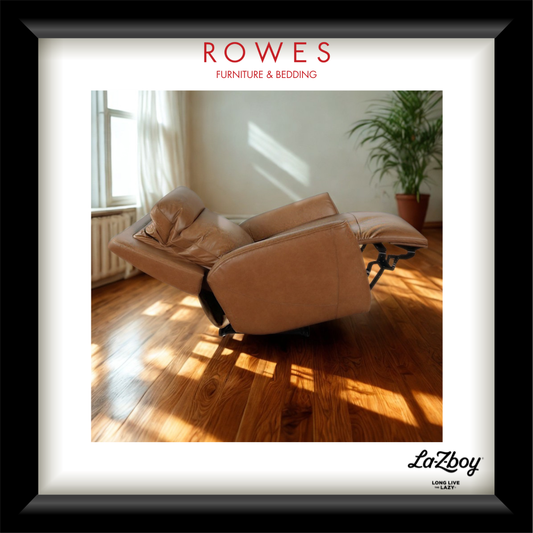 A framed interior photo showing a tan leather recliner chair in a sunlit room with hardwood floors and a potted plant. The frame has a white mat, a black border, the text 'ROWES FURNITURE & BEDDING' at the top, and 'LaZboy' at the bottom right.