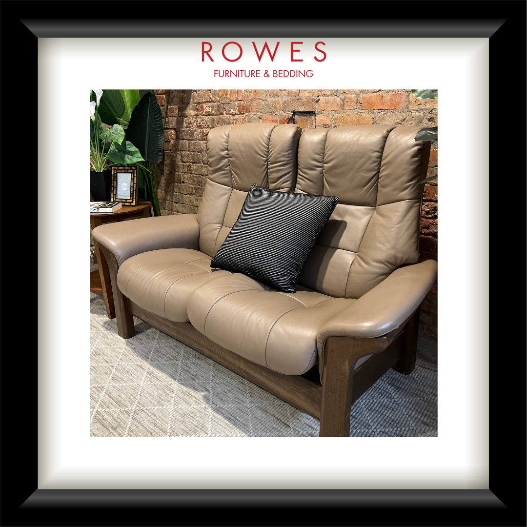 Brown leather sofa with a black pillow in a room with a brick wall and plants, featuring the Rowes Furniture & Bedding logo.
