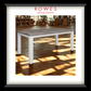 A framed furniture catalog-style print showing a white table with a weathered gray-brown wooden top, set in a warmly lit room with wood floors and shelves. The print is matted with a white border and a black frame, with the text 'ROWES FURNITURE & BEDDOING' at the top.
