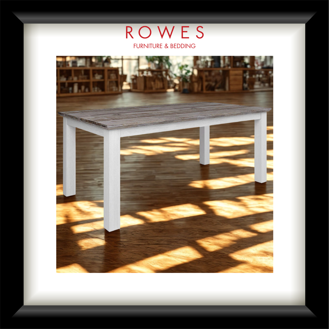 A framed furniture catalog-style print showing a white table with a weathered gray-brown wooden top, set in a warmly lit room with wood floors and shelves. The print is matted with a white border and a black frame, with the text 'ROWES FURNITURE & BEDDOING' at the top.