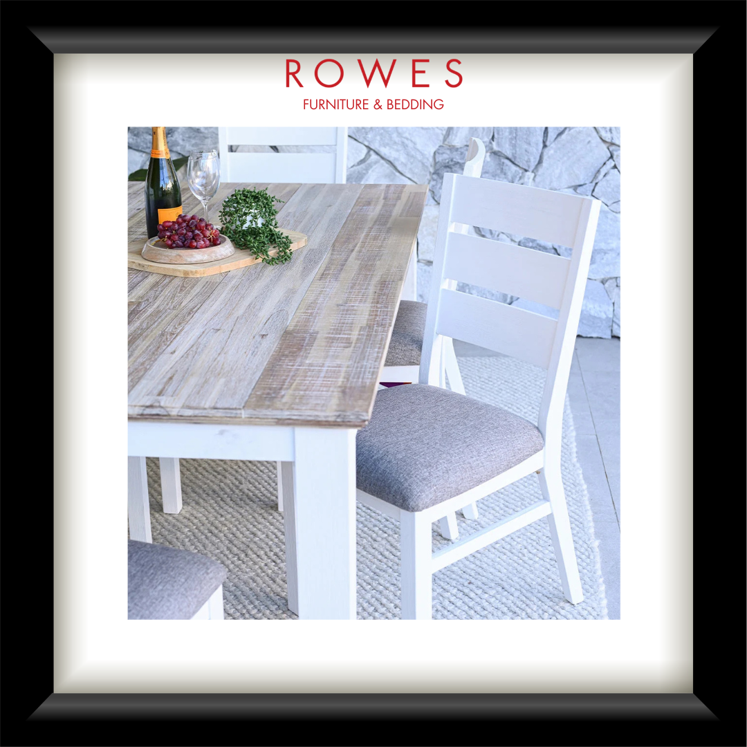 A framed catalog-style interior scene showing a wooden dining table with white chairs, a bottle, grapes, and greenery on a light stone floor, with the text 'ROWES FURNITURE & BEDDING' at the top. The frame is black with a white mat.