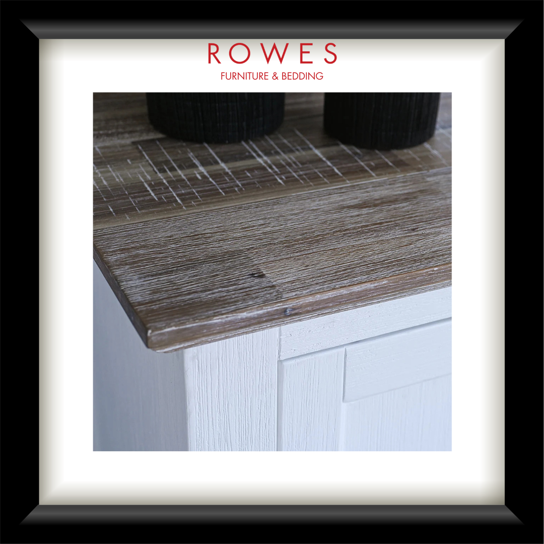 A framed interior photograph showing a rustic weathered wood tabletop corner with white cabinetry below, a white mat, and a black frame. The brand name 'ROWES FURNITURE & BEDDING' appears in red at the top of the frame.