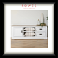 A framed interior photo print featuring a white wooden media console with a light wood top, two open shelves with striped cushions, and two drawers. The print is matted with a wide white border and a thick black frame.