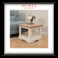 A framed interior photo showing a small square side table with a light natural wood top and white painted frame, placed in a living room beside a pale blue sofa. The frame includes a white mat and a black outer border, with the text 'ROWES FURNITURE & BEDDING' at the top.