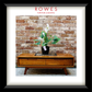 A framed interior photograph showing a mid-century modern wooden console table with two drawers, styled with a potted calla lily on top, set against a brick wall. The image is presented with a white mat and a thick black frame, with the brand name 'ROWES FURNITURE & BEDDING' printed at the top.