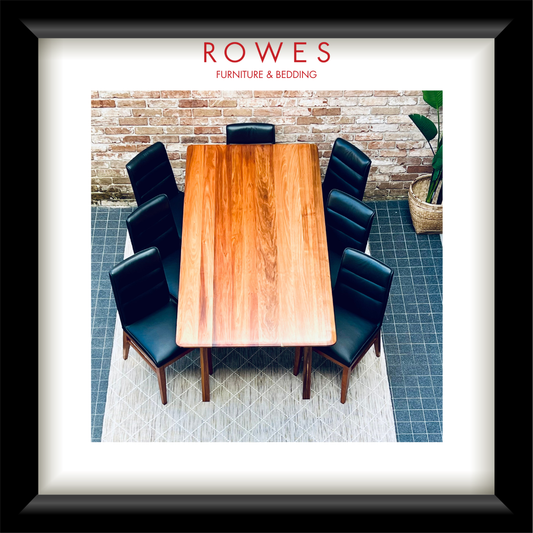 A framed art print featuring a dining room scene with a rectangular wooden table and six black chairs, set against a brick wall and blue tiled floor. The print is matted with a white border and enclosed in a thick black frame.
