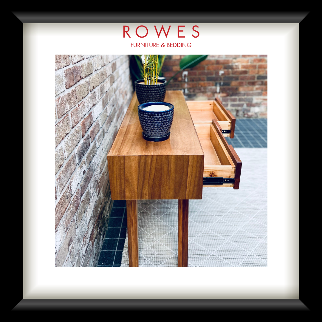 A framed interior design print featuring a wooden console table with drawers, potted plants, and a brick wall, with the text 'ROWES FURNITURE & BEDDING' at the top. The print is matted with a white border and enclosed in a black frame.