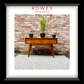 A framed art print featuring a mid-century modern wooden console table against a rustic brick wall, with a potted plant on top and the text 'ROWES FURNITURE & BEDDING' at the top. The print is matted with a white border and enclosed in a thick black frame.