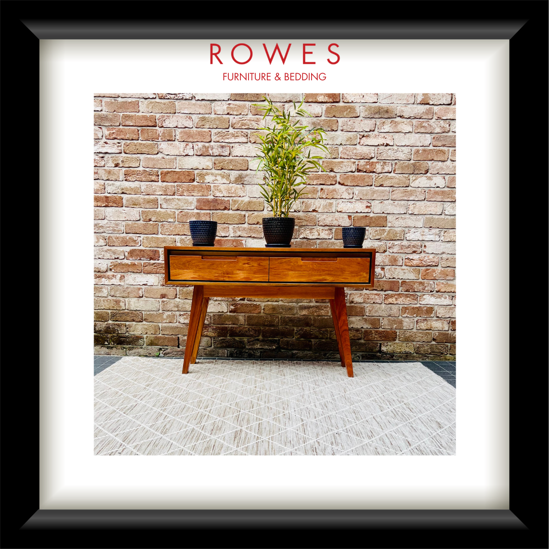 A framed art print featuring a mid-century modern wooden console table against a rustic brick wall, with a potted plant on top and the text 'ROWES FURNITURE & BEDDING' at the top. The print is matted with a white border and enclosed in a thick black frame.