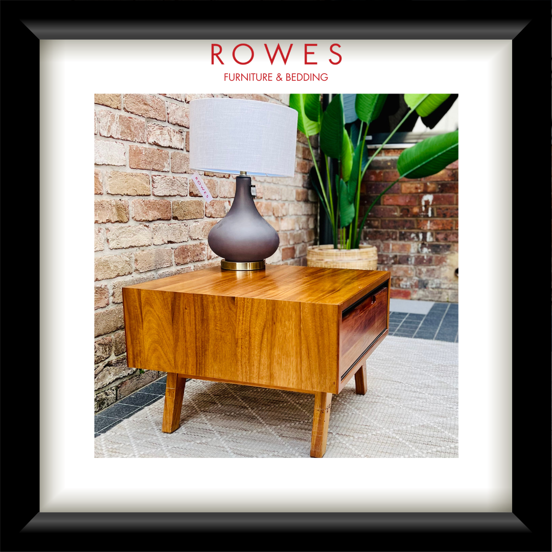 A framed product photo of a mid-century modern wooden side table with a drawer, styled with a table lamp on top. The image is presented with a white mat and a thick black frame, with the brand name 'ROWES FURNITURE & BEDDING' printed at the top.