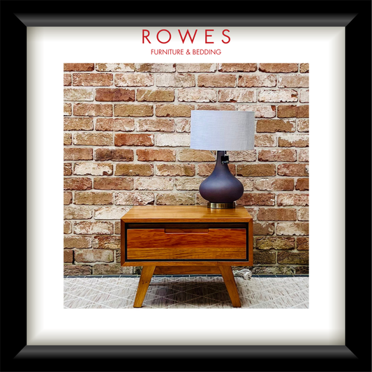 A framed interior design print featuring a mid-century wooden nightstand with a table lamp against a brick wall, with the text 'ROWES FURNITURE & BEDDING' at the top. The print is matted with a white border and enclosed in a black frame.