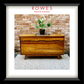 A framed print or poster featuring a mid-century wooden sideboard against a brick wall, with a potted plant on top. The print is matted with a white border and enclosed in a black frame.