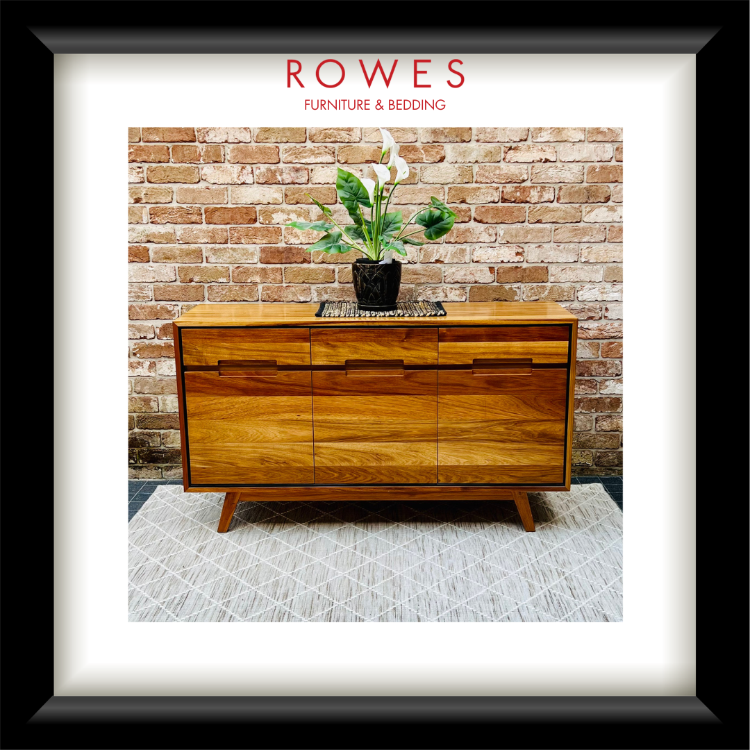 A framed print or poster featuring a mid-century wooden sideboard against a brick wall, with a potted plant on top. The print is matted with a white border and enclosed in a black frame.