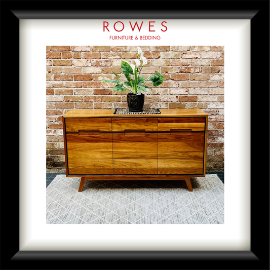 A framed print or poster featuring a mid-century wooden sideboard against a brick wall, with a potted plant on top. The print is matted with a white border and enclosed in a black frame.