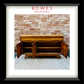 A framed art print featuring a mid-century wooden sideboard cabinet with its doors open, set against a brick wall and patterned rug. The print is matted with a white border and enclosed in a thick black frame.