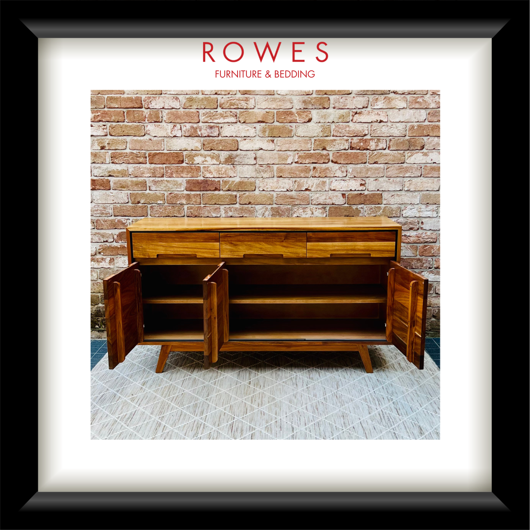 A framed art print featuring a mid-century wooden sideboard cabinet with its doors open, set against a brick wall and patterned rug. The print is matted with a white border and enclosed in a thick black frame.