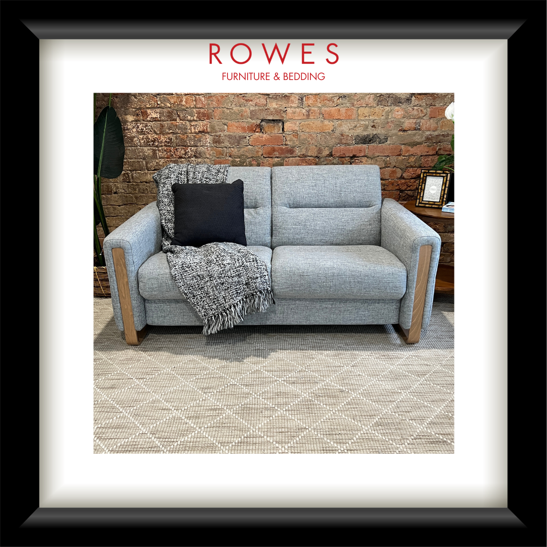 Gray sofa with decorative pillows and a blanket on a rug, against a brick wall with 'Rowes Furniture & Bedding' branding.