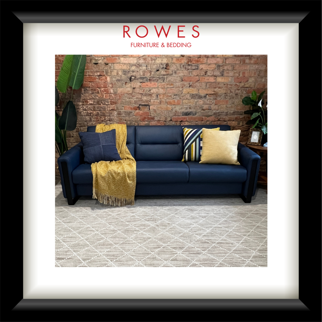 Blue sofa with decorative pillows and a yellow blanket against a brick wall, branded 'Rowes Furniture & Bedding'.
