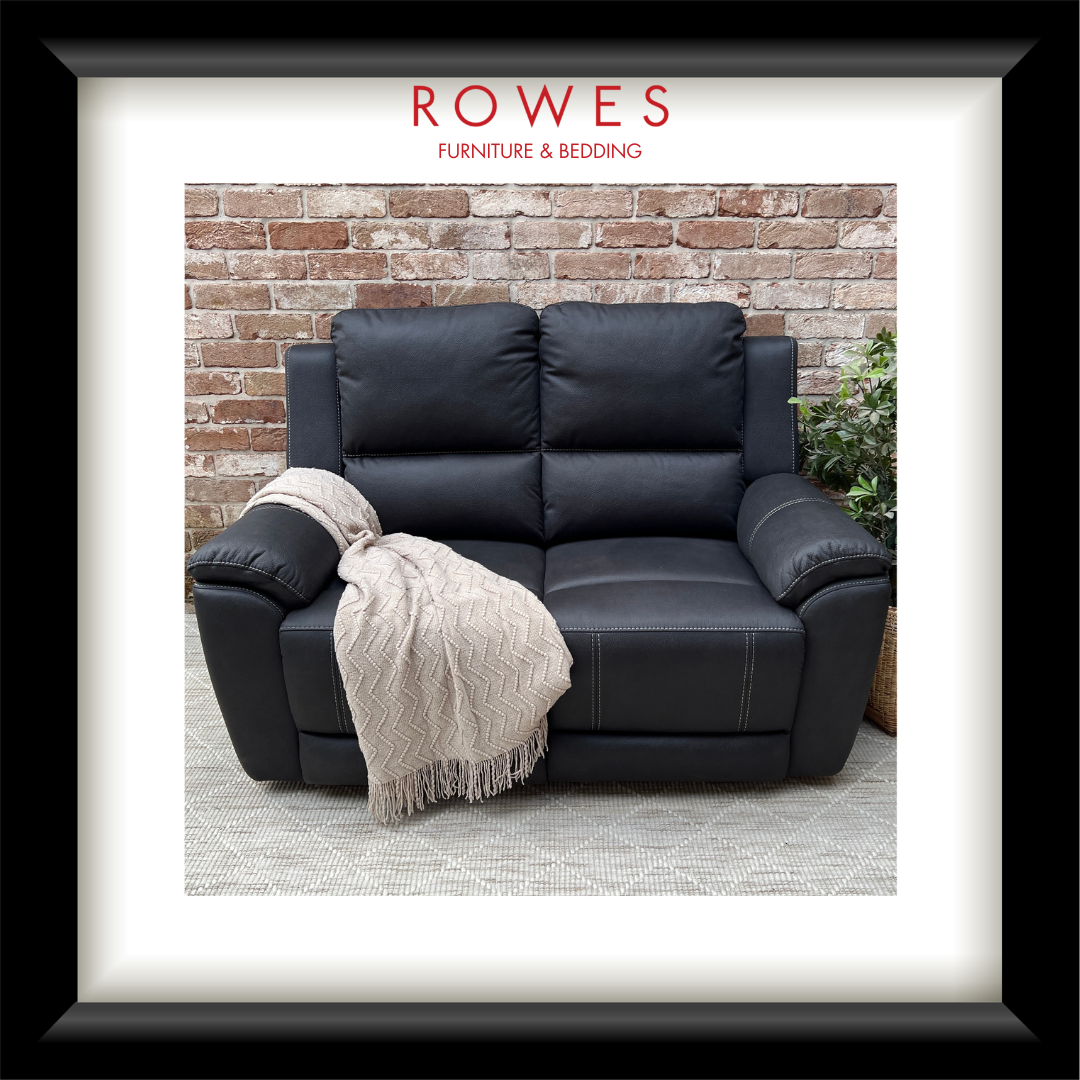 A black leather reclining loveseat shown inside a framed print or matted photo, with a brick wall background and a cream throw blanket draped over one arm. The brand name ROWES FURNITURE & BEDDING appears at the top of the framed presentation.