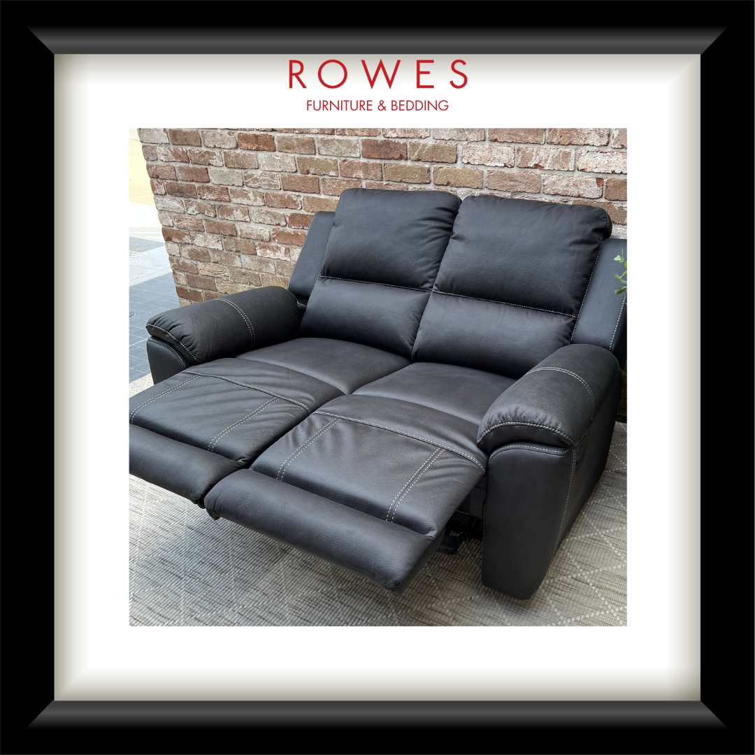 A black leather reclining loveseat sofa shown in a showroom setting with a brick wall background, framed by a white mat and a black frame. The brand name 'ROWES FURNITURE & BEDDING' is printed at the top of the image.