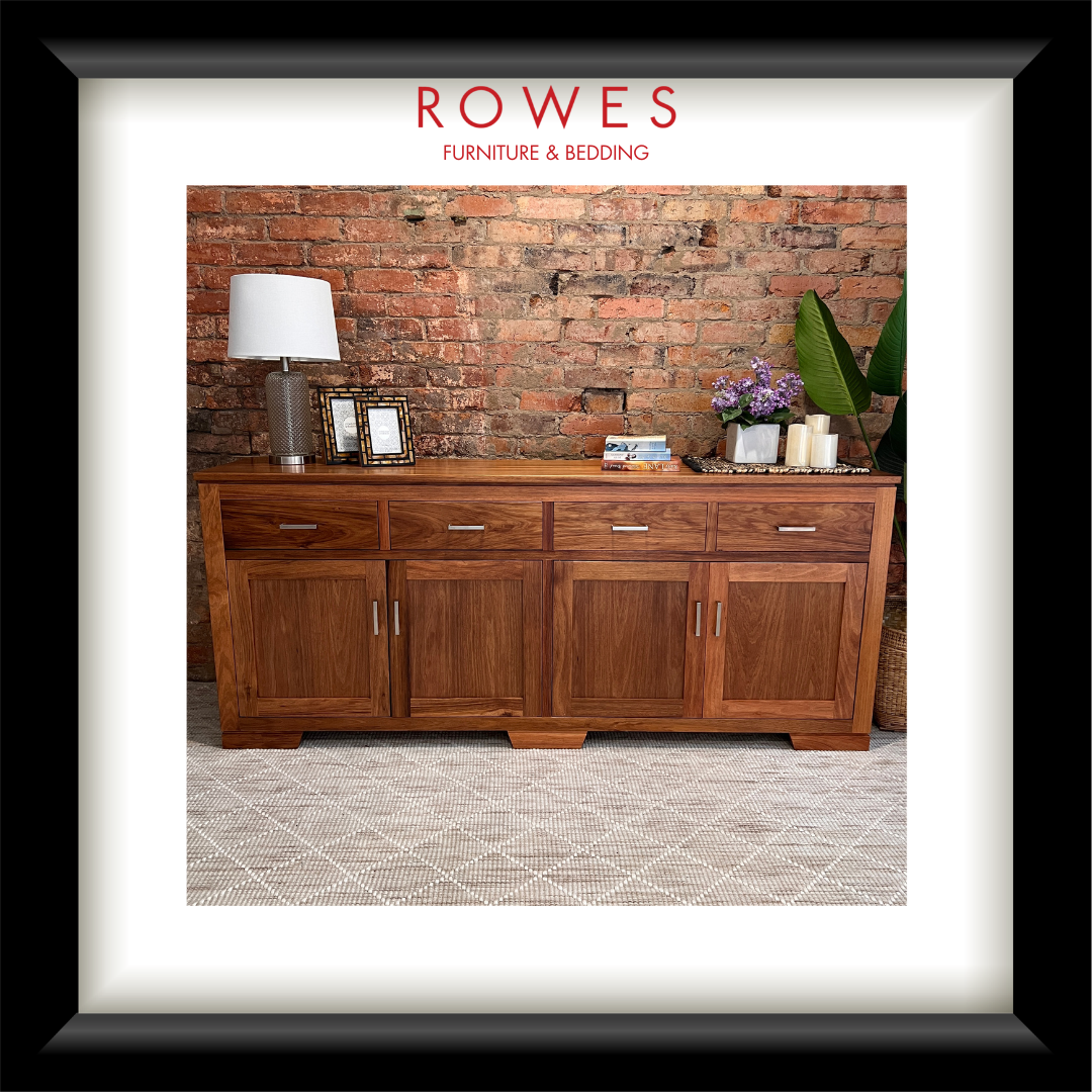 A framed interior photo print featuring a wooden sideboard or credenza in a warm medium brown finish, styled with a lamp, picture frames, candles, and flowers against a brick wall. The print is matted with a white border and enclosed in a thick black frame.