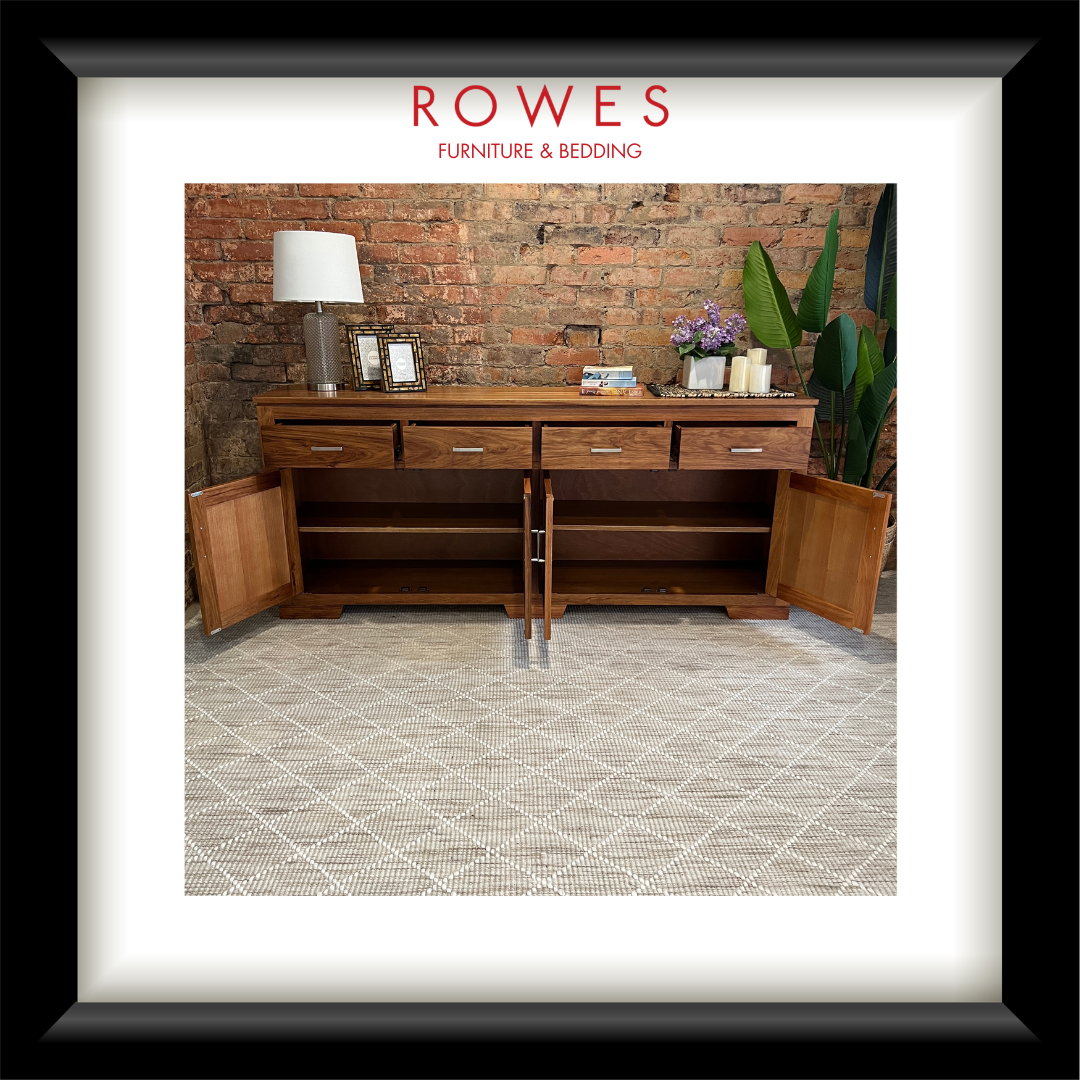 A framed interior photo print featuring a wooden sideboard or credenza in a warm medium brown finish, styled with a lamp, picture frames, candles, and flowers against a brick wall. The print is matted with a white border and enclosed in a thick black frame.
