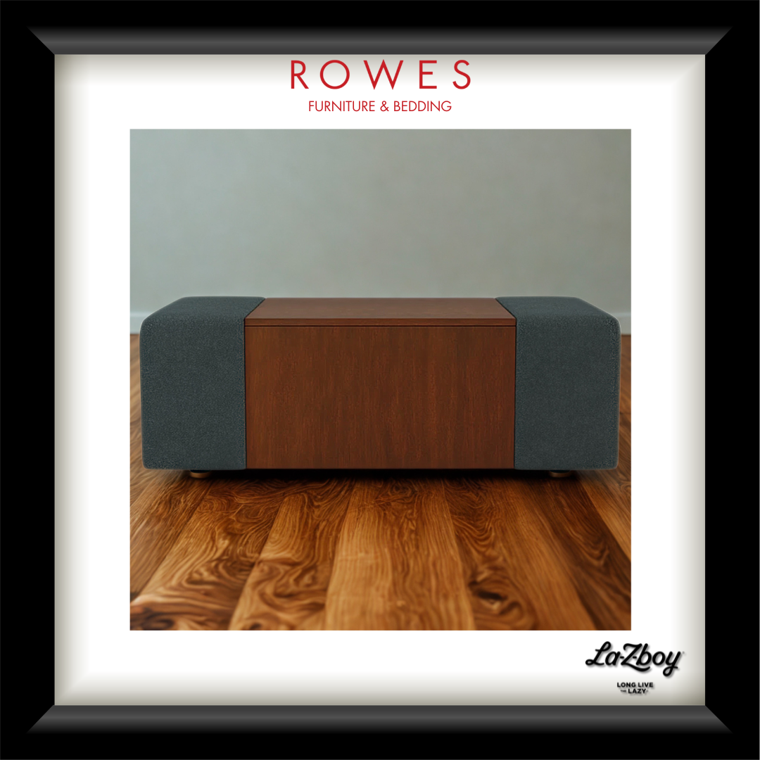 A framed product photograph of a modern rectangular ottoman or coffee table with a dark wood center panel and dark gray fabric side panels, set on a wood floor against a light gray wall. The frame has a white mat and a black frame, with the brand name 'ROWES FURNITURE & BEDDING' at the top and 'LaZboy' at the bottom right.