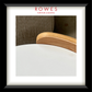 A framed interior design print or magazine-style page with a black frame, white mat, and the visible brand text 'ROWES FURNITURE & BEDDOING' at the top. The central photo shows part of a light wood chair back and a white tabletop against a neutral indoor background.