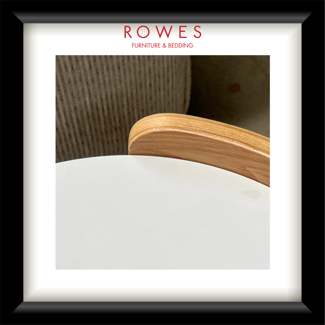 A framed interior design print or magazine-style page with a black frame, white mat, and the visible brand text 'ROWES FURNITURE & BEDDOING' at the top. The central photo shows part of a light wood chair back and a white tabletop against a neutral indoor background.