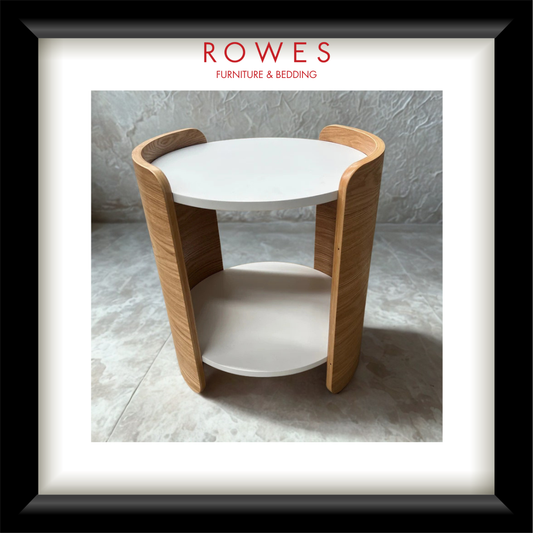 A framed product photograph of a modern round side table with a white top and shelf, supported by curved light oak wood legs, presented with a white mat and black frame. The brand name 'ROWES FURNITURE & BEDDING' is printed at the top of the photo.