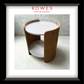 A framed catalog-style presentation of a modern cylindrical side table with a white round tabletop and curved light wood side panels, set against a light gray interior backdrop. The frame includes the text 'ROWES FURNITURE & BEDDING' at the top.