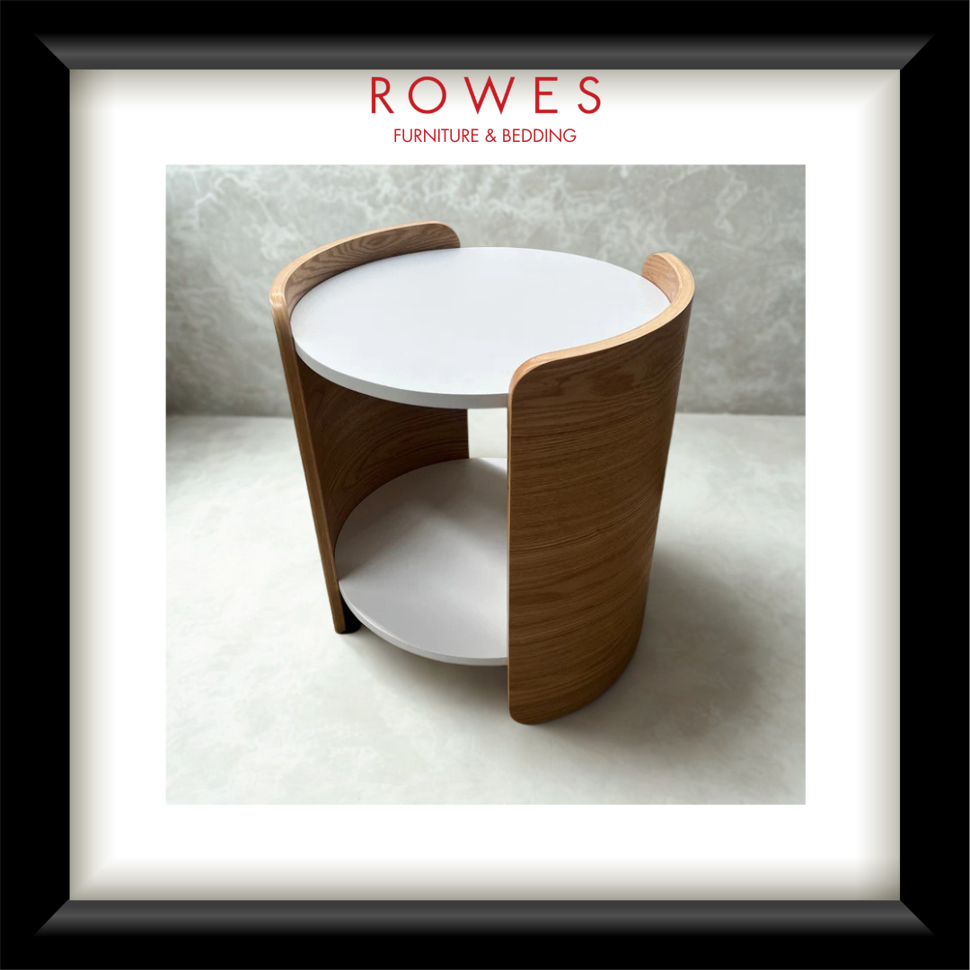 A framed catalog-style presentation of a modern cylindrical side table with a white round tabletop and curved light wood side panels, set against a light gray interior backdrop. The frame includes the text 'ROWES FURNITURE & BEDDING' at the top.