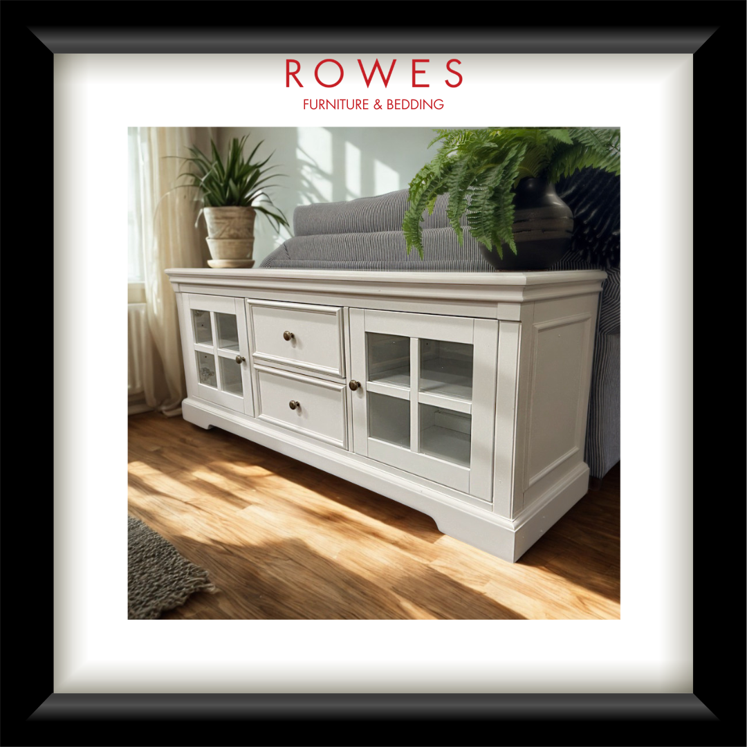 A framed catalog-style image showing a white wooden storage cabinet with glass-front doors and drawers in a living room setting. The top of the frame includes the text 'ROWES FURNITURE & BEDDING' above the photo, with a black outer frame and white mat.