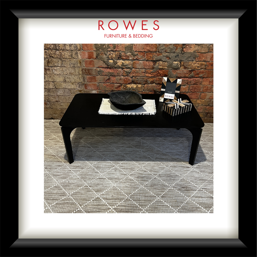 A framed image of a black coffee table on a beige rug, set against a brick wall. On the coffee table sits ornaments and a photo frame. The Rowes logo sits at the top of screen.