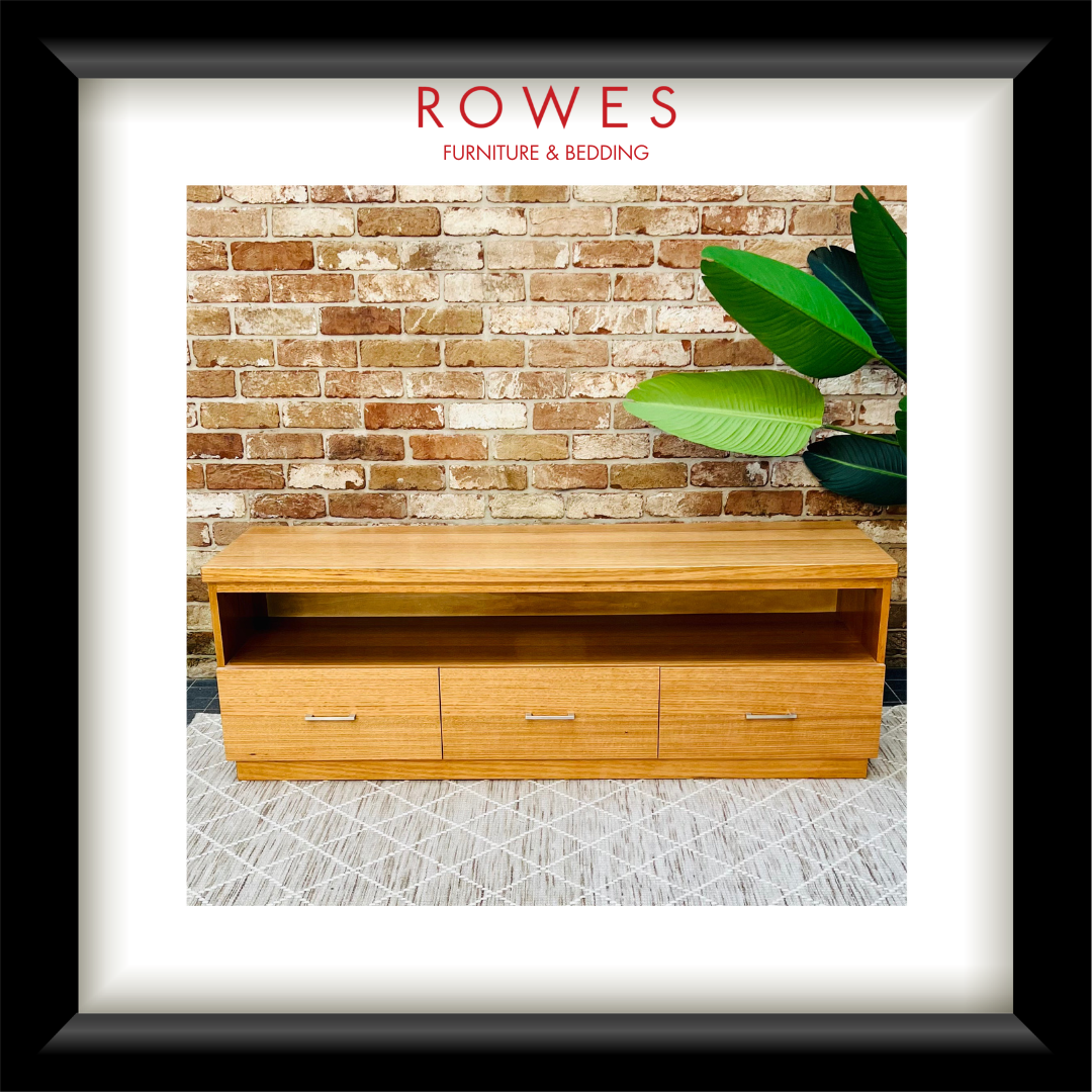 A framed product photo of a light wood bench or low cabinet with three drawers and open shelving, set against a brick wall with a green plant. The image is matted with a white border and a black frame, with the text 'ROWES FURNITURE & BEDDING' at the top.