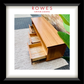 A framed product photo of a light wood bench or low cabinet with three drawers and open shelving, set against a brick wall with a green plant. The image is matted with a white border and a black frame, with the text 'ROWES FURNITURE & BEDDING' at the top.