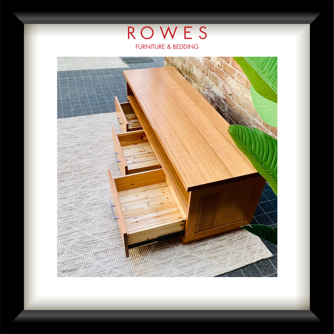 A framed product photo of a light wood bench or low cabinet with three drawers and open shelving, set against a brick wall with a green plant. The image is matted with a white border and a black frame, with the text 'ROWES FURNITURE & BEDDING' at the top.
