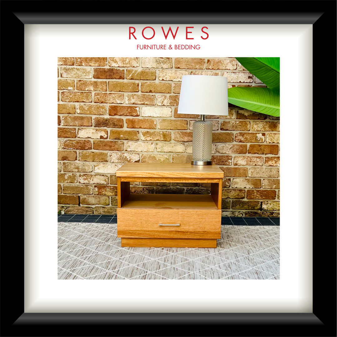 A framed product photograph of a light wood nightstand with a drawer, styled with a table lamp against a brick wall and patterned rug. The image is presented with a white mat and a thick black frame, with the brand name 'ROWES FURNITURE & BEDDING' printed at the top.