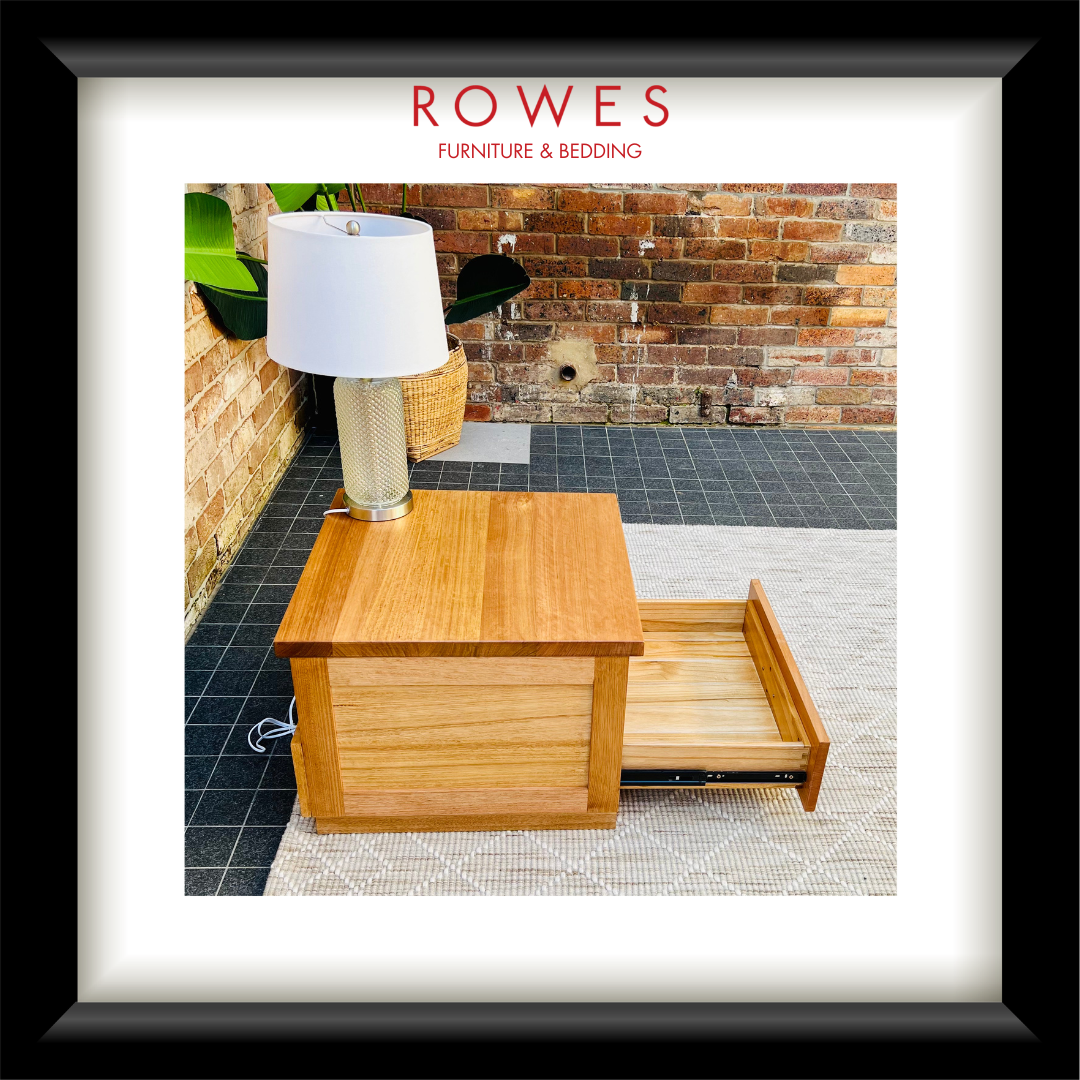 A framed product photo of a wooden side table or nightstand with one drawer pulled open, shown on a patio with a white lamp on top. The image is matted with a white border and a black frame, with the text 'ROWES FURNITURE & BEDDING' at the top.